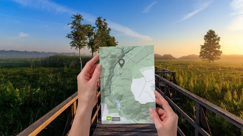 Person hält eine gedruckte Karte mit Standortmarkierung in einer Landschaft auf einem Holzsteg